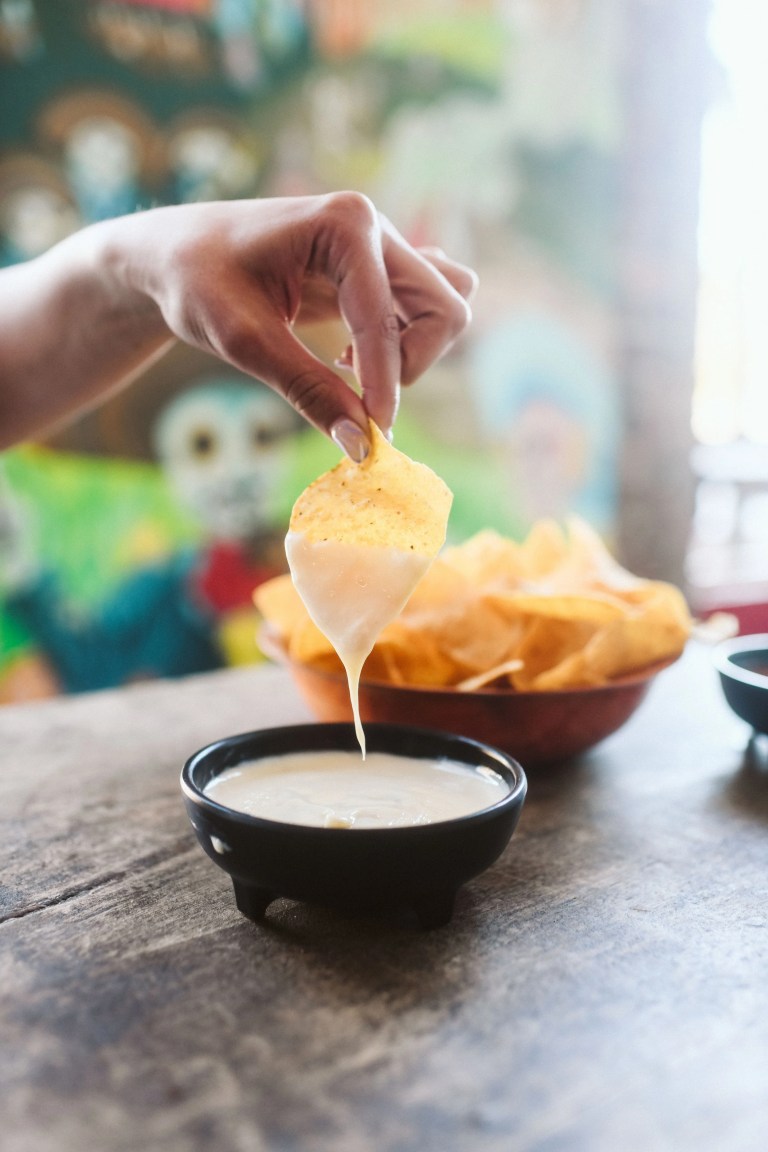 Hand Holding Tortilla Chip Dipped in Queso Sauce 768x1152